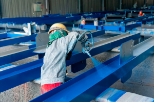 Imagem de Vinilak Pintor industrial com EPI aplica tinta dupla função Vinilak com pistola em estrutura metálica azul. O jato de tinta líquida cria uma névoa visível sobre o aço.